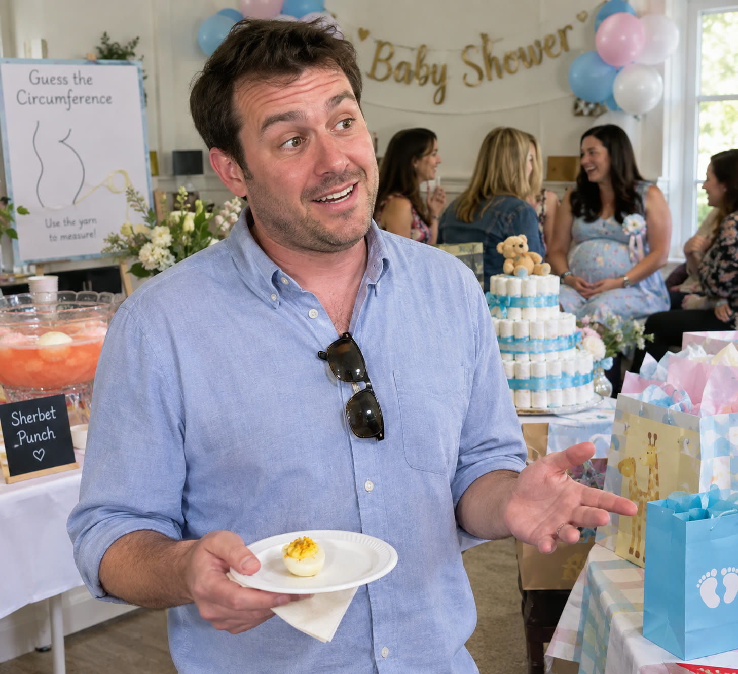A man in a blue button-down shirt holds a paper plate with a single deviled egg at a baby shower, gesturing mid-sentence in front of a sherbet punch bowl, a diaper cake and a “Guess the Circumference” yarn game sign.