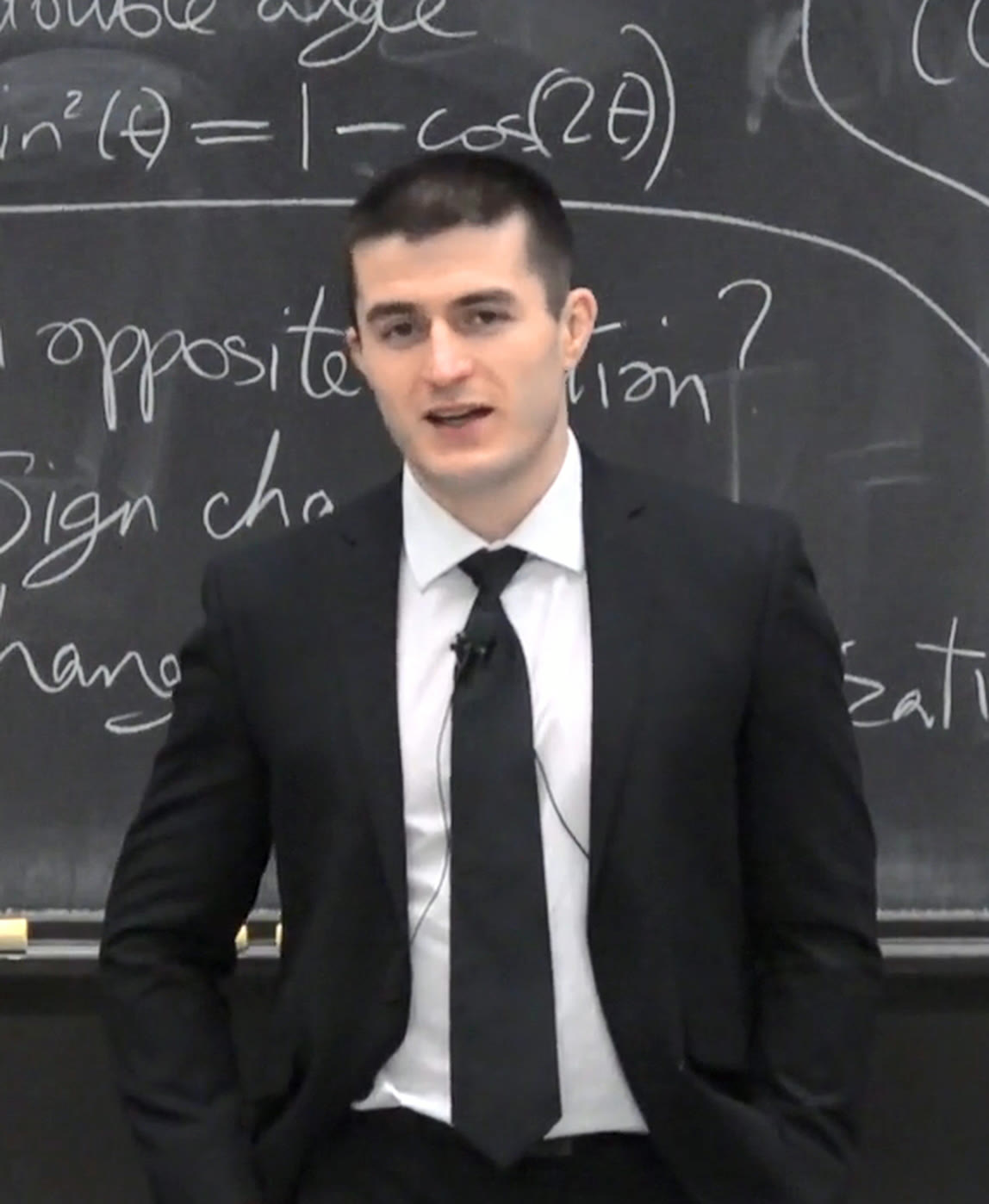 Lex Fridman in a dark suit and tie, teaching in front of a blackboard covered in trigonometric identities.