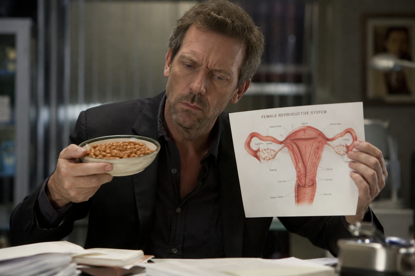 A man in a dark jacket with stubble sits at a cluttered desk holding up a bowl of baked beans in one hand and a printed anatomical diagram headed “Female Reproductive System” in the other, looking at the beans with a sceptical frown.