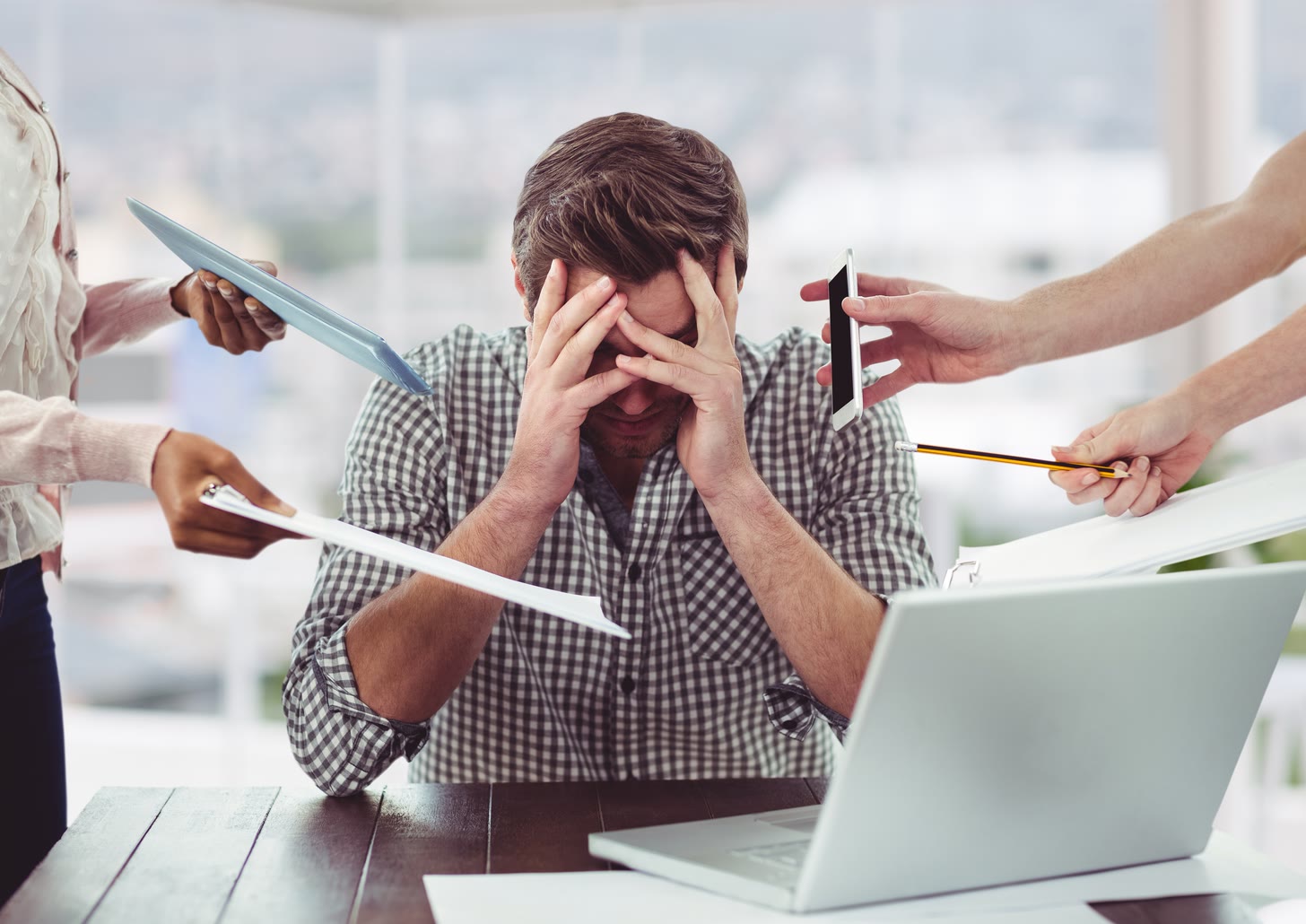 A man at a desk holds his head in both hands as four coworkers reach in from all sides holding a tablet, a clipboard, a phone and a pencil.