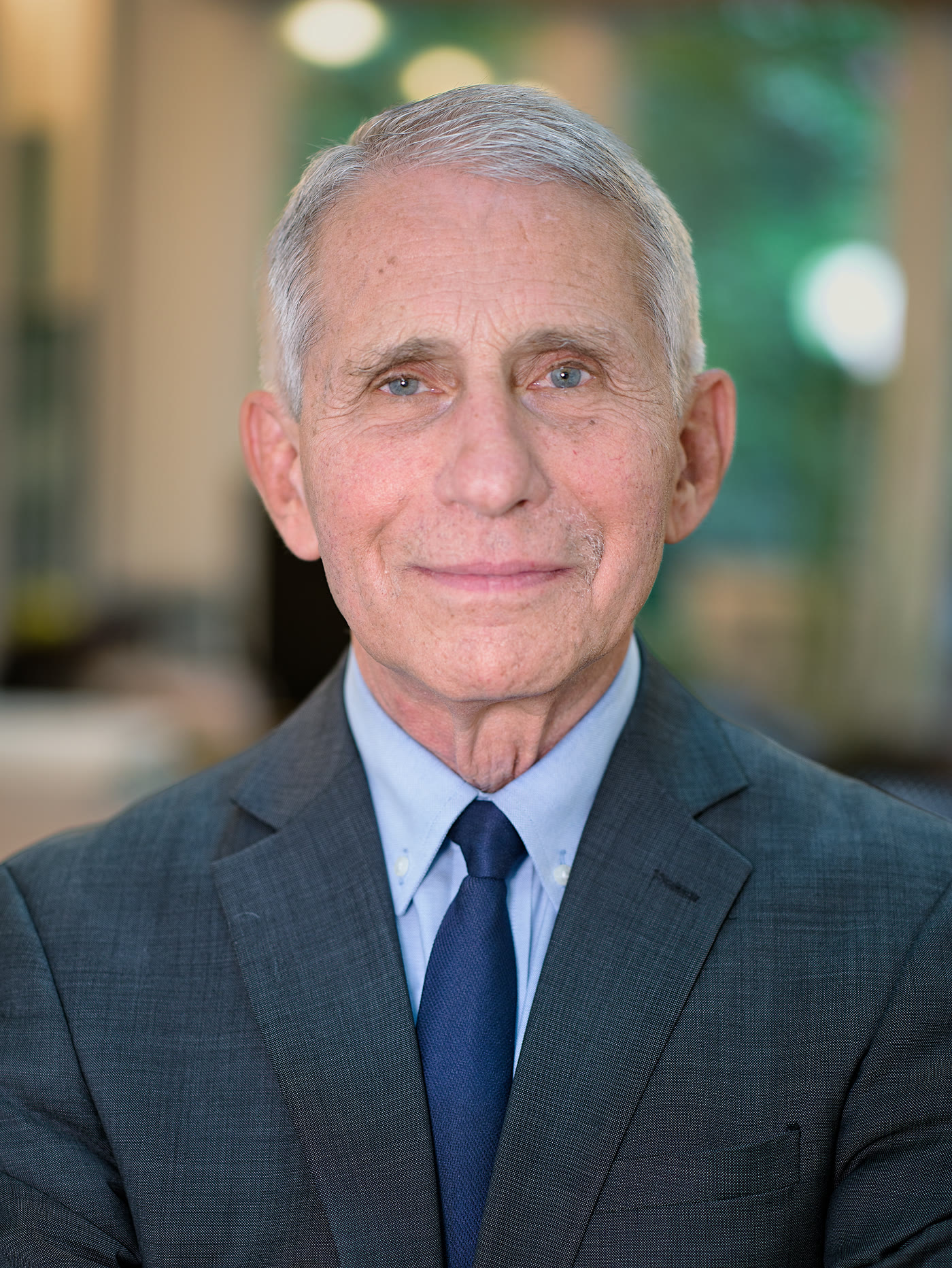 Portrait photograph of Anthony Fauci in a dark suit and blue tie, photographed indoors against a softly blurred background.