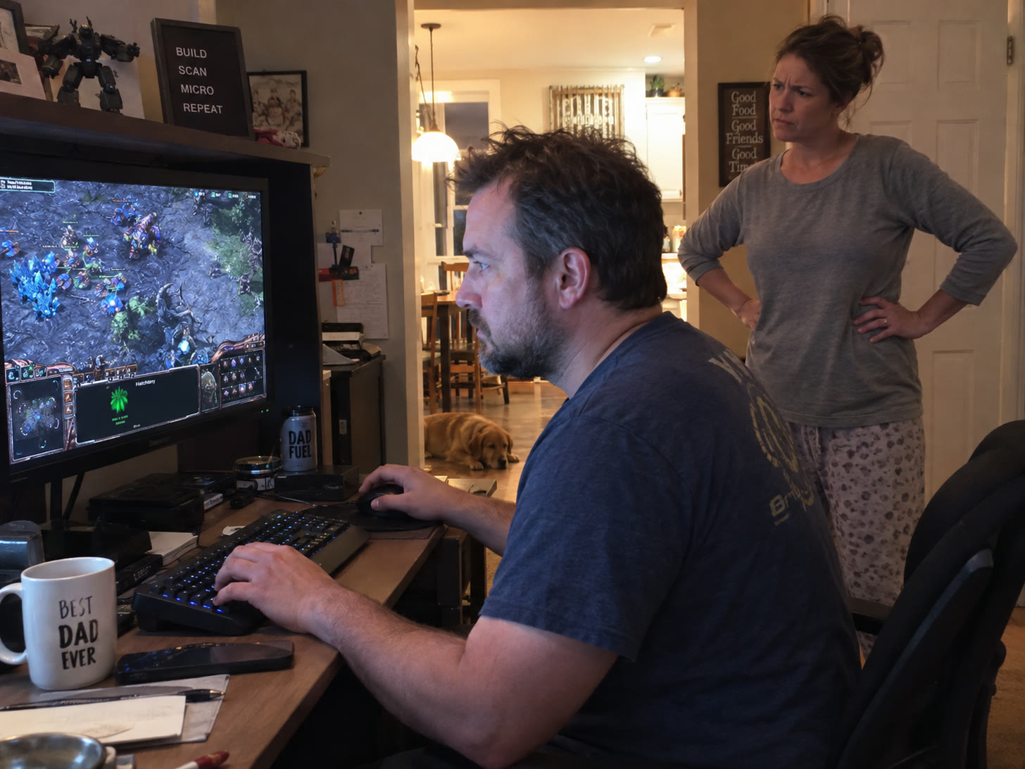 A man in his forties sits at a desk in a family room playing a real-time strategy game on a large monitor, one hand on the mouse and one on a keyboard. A woman in pyjamas stands in the doorway behind him with her hands on her hips, watching. A lettered board above the monitor reads BUILD, SCAN, MICRO, REPEAT. A mug on the desk reads BEST DAD EVER and a bottle beside the monitor reads DAD FUEL. A dog lies on the floor of the hallway behind them.