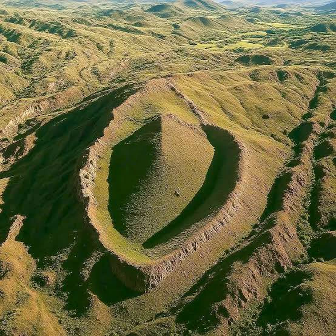 An aerial photograph of an elongated, boat-shaped raised formation on a green hillside, with a raised outer ridge enclosing a smooth central area.