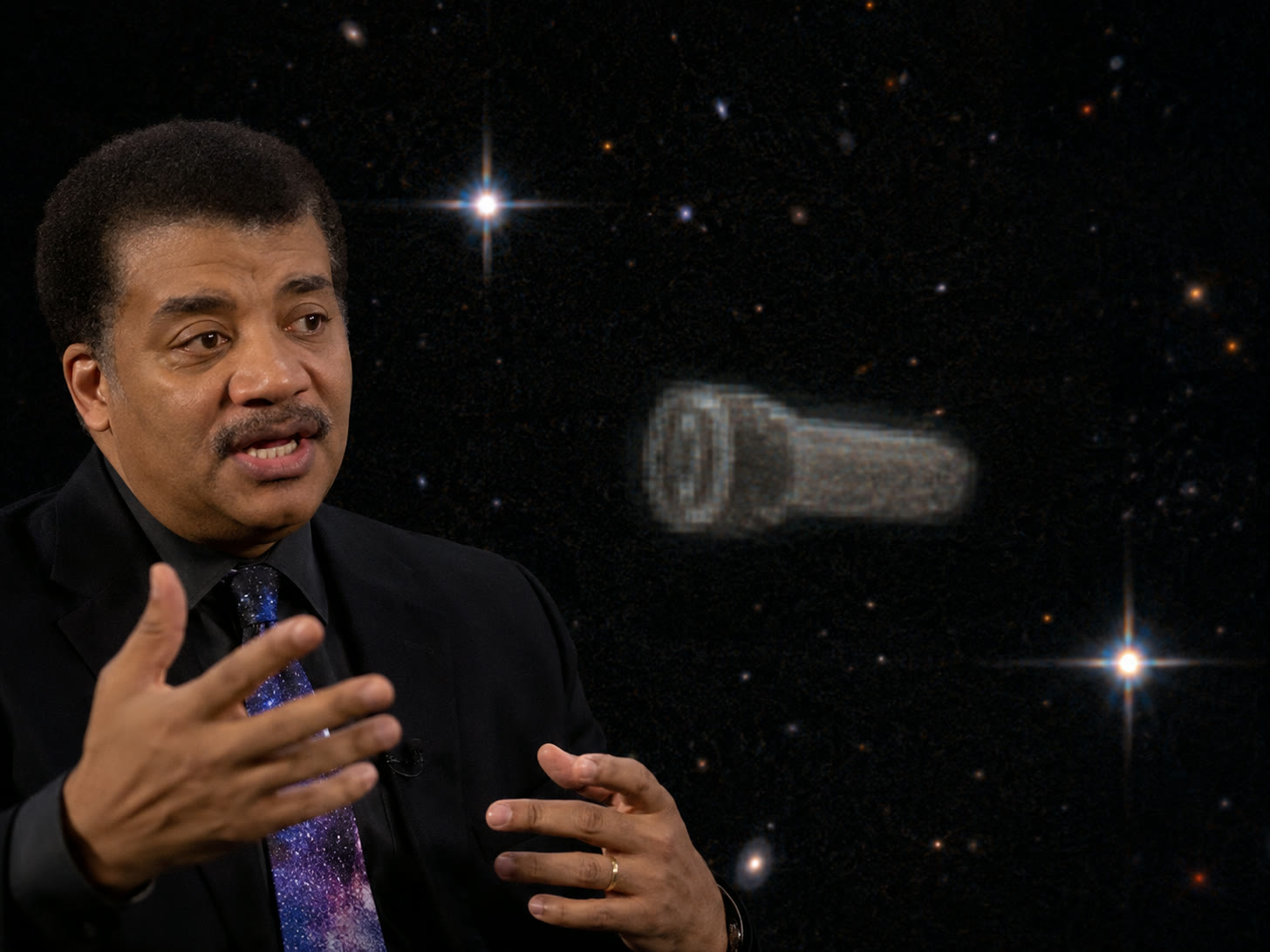 A composite image: the astrophysicist Neil deGrasse Tyson in a dark suit and galaxy-patterned tie, gesturing mid-sentence, with a grey cylindrical object rendered against a starfield behind him.